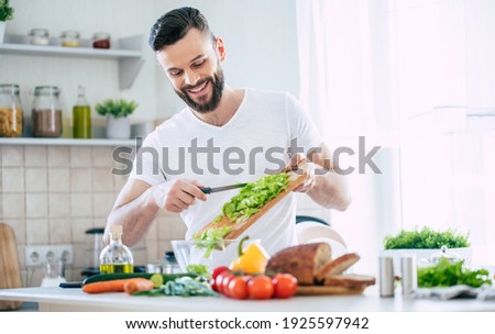 Handsome happy bearded man is preparing wonderful fresh vegan salad in the kitchen at home