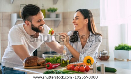 Excited smiling young couple in love making a super healthy vegan salad with many vegetables in the kitchen and man testing it from a girl's hands Royalty-Free Stock Photo #1925598080