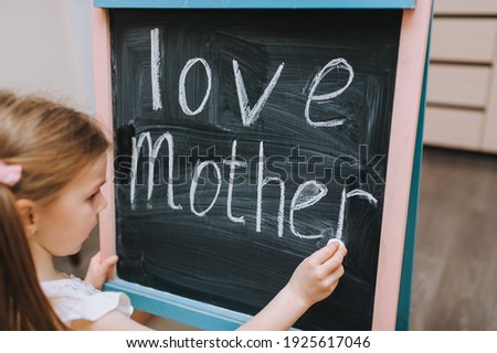 Red-haired little girl painstakingly writes on a wooden black board, easel, holding white chalk in her hand, the words love and mom. Photography, copy space, concept.