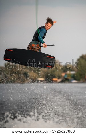 healthy athletic guy in wetsuit and vest skillfully jumps on wakeboard above the splashing river wave