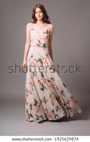 Candid portrait of a brunette young woman in summer gown with shoulder straps and floral print. Calm studio portrait of young lady in long sleeveless evening gown on grey background. Royalty-Free Stock Photo #1925629874