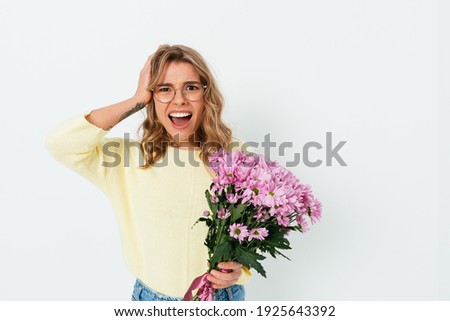 Unhappy young woman in glasses and casual clothes holding bouquet of flowers and clutching her head with puzzled facial expression on white studio background.