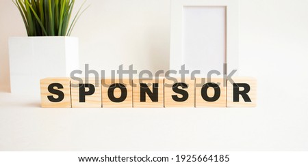 Wooden cubes with letters on a white table. The word is SPONSOR. White background with photo frame, house plant.