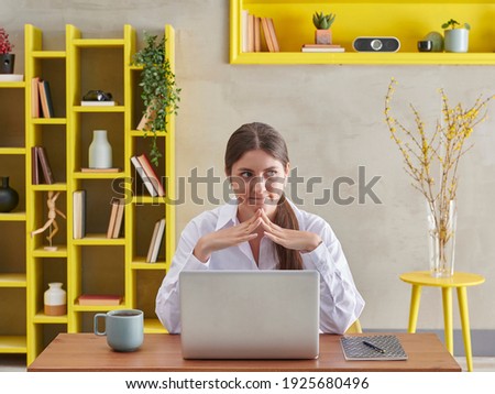 Woman in white shirt is is thinking in video talking, grey wall background and yellow bookshelf, coffee laptop style.