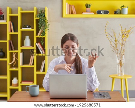 Woman in white shirt is asking a question in video talking, grey wall background and yellow bookshelf, coffee laptop style.
