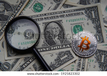 Pile of US dollar cash. Next to it are a number of gold bitcoin digital cryptocurrency coins and a magnifying glass. Bank image and photo background. 