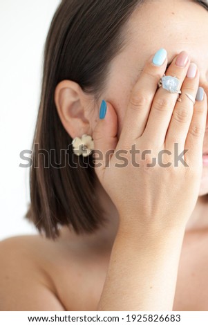 Cropped photo of a woman with short dark hair in white clothes on white background. Portrait of a female covering her face with her hand. Girl has two rings big blue stone on the fingers.