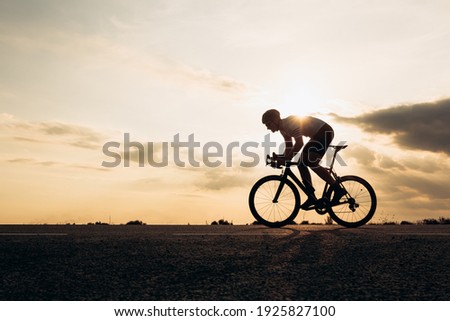 Side view of active sportsman in protective helmet riding bike among nature during sunset. Muscular guy in activewear cycling on paved road. Royalty-Free Stock Photo #1925827100