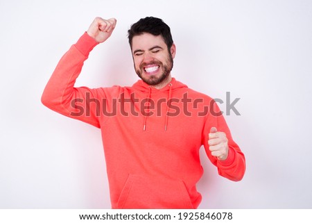 Attractive Young Caucasian bearded man wearing pink hoodie against white background celebrating a victory punching the air with his fists and a beaming toothy smile.