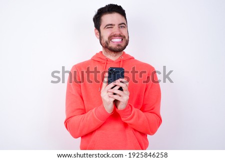 Pleased young Caucasian bearded man wearing pink hoodie against white background using self phone and looking and winking at the camera. Flirt and coquettish concept.