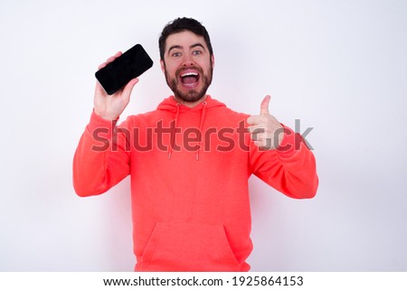 Portrait young Caucasian bearded man wearing pink hoodie against white background holding in hands cell showing giving black screen thumb up