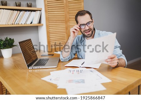 Shocked amazed perplexed young bearded business man 20s wearing blue shirt glasses sitting at desk hold looking on papers document keeping mouth open working on laptop pc computer at home or office