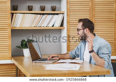 Handsome smiling young bearded business man in blue shirt glasses sitting at desk with papers document working on laptop pc computer making video call waving ang greeting with hand at home or office