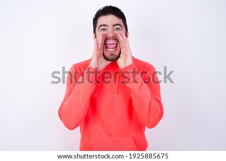 Young Caucasian bearded man wearing pink hoodie against white background shouting excited to front.