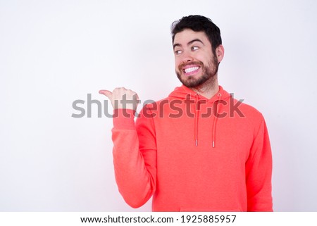 Charming Young Caucasian bearded man wearing pink hoodie against white background looking at copy space having advertisements