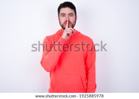 Young Caucasian bearded man wearing pink hoodie against white background  makes silence gesture, keeps finger over lips. Silence and secret concept.