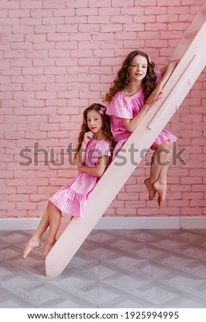 Portrait of girls with long curled hair in a pink dress on a pink background. They sit on the stairs on different steps.