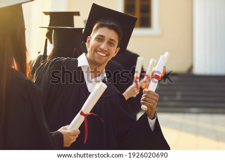 Young happy university graduate in a mantle demonstrates his diploma and smiles at the camera next to his classmates. Concept of success and goal achievement. Royalty-Free Stock Photo #1926020690