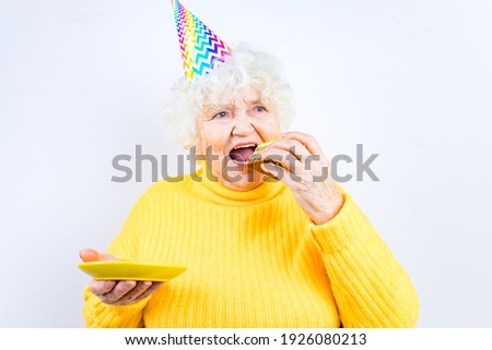 older woman with a gift wear yellow sweater and horn cap on a white background holding plate with cake