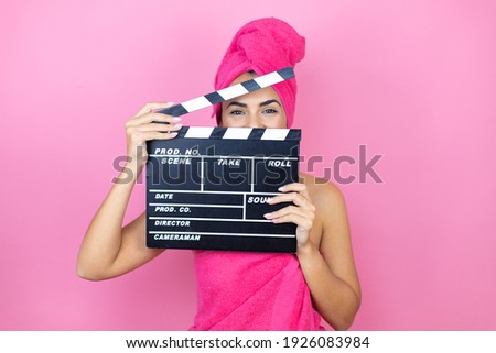 Young beautiful woman wearing shower towel after bath standing over isolated pink background holding clapperboard very happy having fun