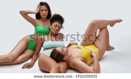 Three multiethnic young female models with different body types in colorful underwear posing for camera, lying on the floor isolated over white background. Friendship, beauty, body positive concept