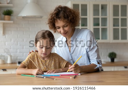 Developing creativity. Happy young foster mother play with little adopted daughter paint in album with colored pencils. Caring millennial nanny help small girl to draw teach child to create picture