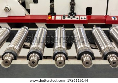 Lined up magnetic cylinders for die cut on rotary printing press. Magnetic cylinder for flexo rotary die cutting. Magnetic roll and in-line press machine in background. Cylinder for cutting dies. Royalty-Free Stock Photo #1926116144