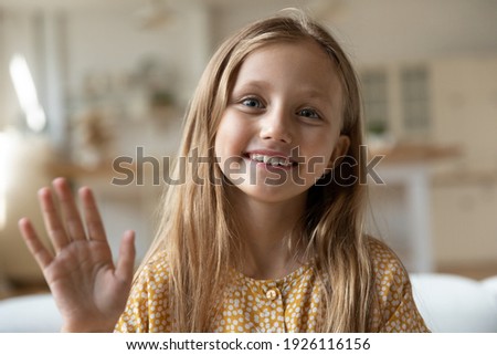 Hello, mommy. Headshot portrait of adorable little junior girl calling parent from home by video link looking at camera with smile. Web cam view of cute 7 year old kid greet friend online wave hand hi