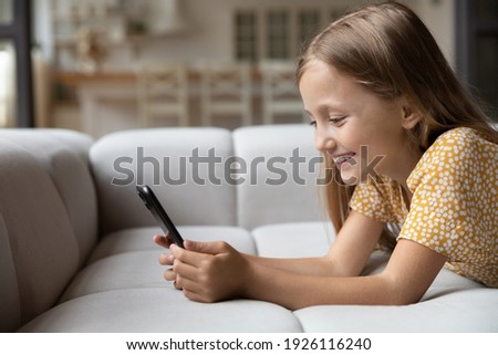 Smiling little child lying on soft couch at living room look at phone screen use game app contact mates at social network. Funny small girl engaged in watching online channel for children. Copy space