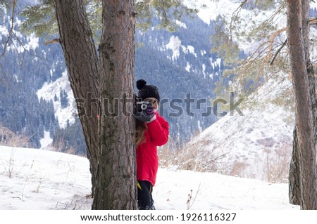 A young girl with blond hair and a red jacket, photographing a mountain landscape, trekking in the mountains and taking pictures with a camera. Photographer girl in the mountains among the trees