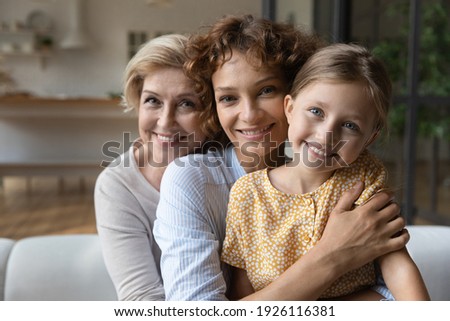 Caring grown woman mother hold on knees hug girl kid while mature female grandmother pensioner embrace her adult daughter from back. Family portrait of women dynasty look at camera sit on sofa cuddle