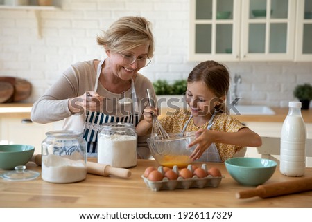 Cooking with soul. Happy little girl in apron help senior grandma at kitchen mix dough for cookies pancakes. Smiling older granny teach small grandkid to bake homemade cake pastry share family recipe Royalty-Free Stock Photo #1926117329