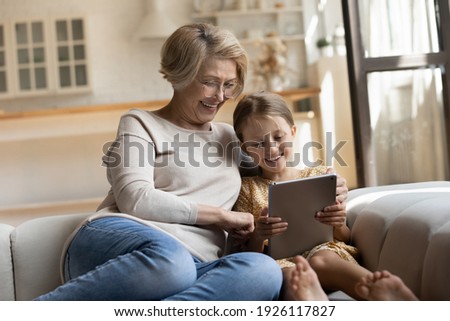 Having fun. Smiling retired woman nanny sit on cozy couch embrace little girl watch funny cartoon at web online using tablet. Bonding senior granny and small grandchild enjoy video clip on digital pad