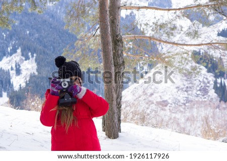 A young girl with blond hair and a red jacket, photographing a mountain landscape, trekking in the mountains and taking pictures with a camera. Photographer girl in the mountains among the trees
