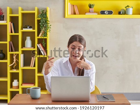 Woman in white shirt is confirm in front of the laptop, video talking, grey wall background and yellow bookshelf, coffee laptop style.