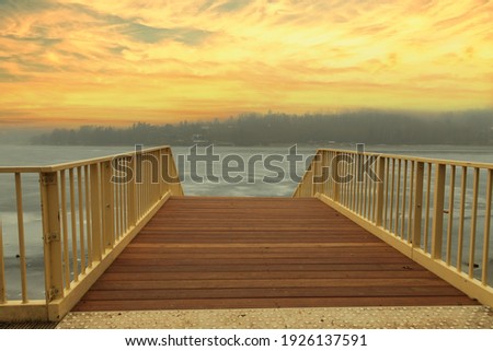 A metal pier with a wooden floor leads to the water. Pier for the steamer Sokolak in the city of Brno in the Czech Republic. There is ice on the water. Sky at sunset.