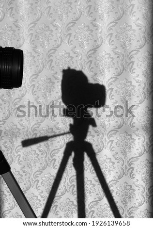 Shadow of old SLR film camera on tripod on wall. In foreground are fragments of camera lens and stand. Working moment in photo studio. Focus on background. Monochrome. Vertical photo.
