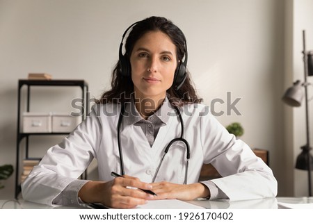 Head shot portrait confident female doctor wearing headphones and uniform with glasses taking notes, looking at camera, therapist physician consulting patient, video call, telemedicine concept Royalty-Free Stock Photo #1926170402