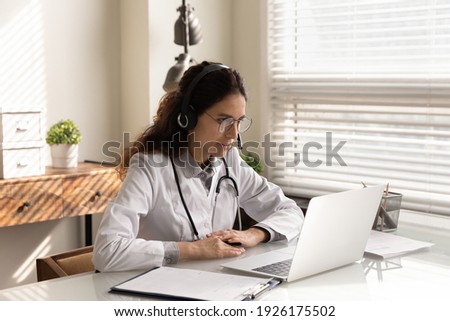 Professional female doctor physician wearing headphones and uniform with glasses consulting patient client online by video call, talking, looking at laptop screen, telemedicine service concept Royalty-Free Stock Photo #1926175502