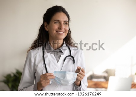 Head shot close up smiling professional female doctor holding medical protective face mask, physician therapist wearing uniform looking to aside, thinking about future, enjoying break in hospital Royalty-Free Stock Photo #1926176249