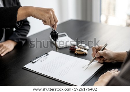 The renter is signing a car rental agreement with the car rental company. After discussing the details and charges with the employee, the employee hand over the car keys to the renter. Royalty-Free Stock Photo #1926176402