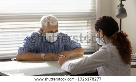 Close up female doctor consulting smiling older patient wearing medical face mask about health insurance agreement at meeting in hospital, therapist and elderly patient discussing contract terms