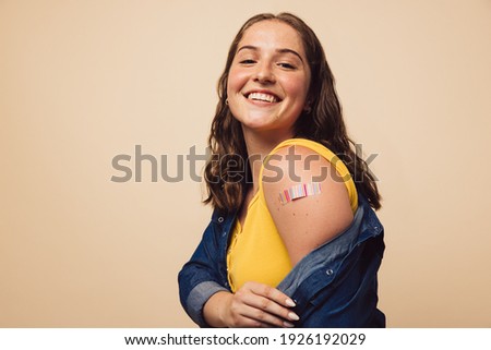 Portrait of a female smiling after getting a vaccine. Woman holding down her shirt sleeve and showing her arm with bandage after receiving vaccination. Royalty-Free Stock Photo #1926192029