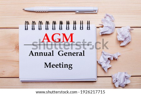 On a wooden background, a white notebook with the inscription AGM Annual General Meeting, a white pen and crumpled white pieces of paper