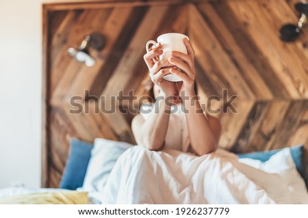 Lazy female lying down under the white blanket on the linen bed and holding the fresh coffee cup in the early morning. Lazy day off and "coffee in bed"concept image. 