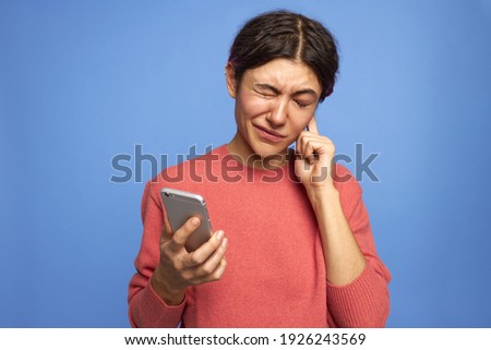 Isolated shot of unhappy student girl with nose ring rubbing ear having hearing problem holding generic mobile phone watching lecture online on video conference chat. Technology and communication