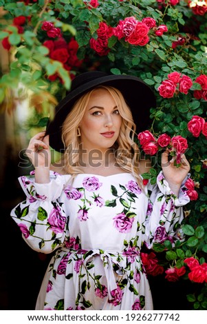 Outdoor photo of romantic young woman in rose garden. girl with red lips in a dress with a print of roses. Young model in hat on a background of a bush of roses. Stylish woman.