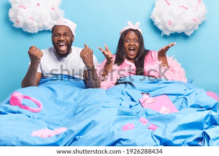 Positive dark skinned man clenches fists and exclaims loudly poses near his puzzled Afro American wife in bed. Emotional couple react on amazing news isolated over blue background. Home lifestyle