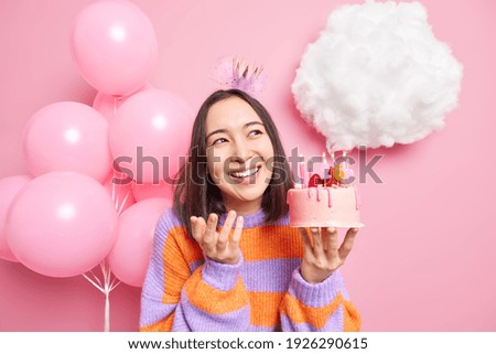 Indoor shot og happy pleasant looking woman smiles broadly holds tasty cake with burning number candles celebrates birthday on party poses against balloons has festive mood. Holidays concept