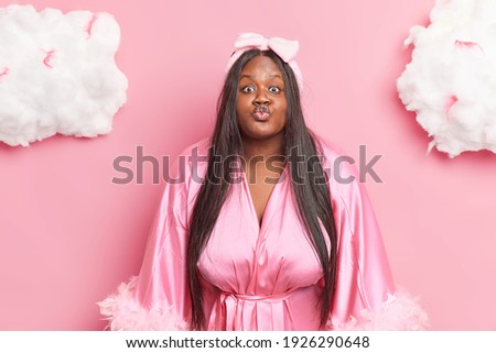 Romantic plump long haired beautiful woman folds lips at mwah want to kiss passionately someone wears domestic robe poses isolated over pink background white cluds above. Women domestic style